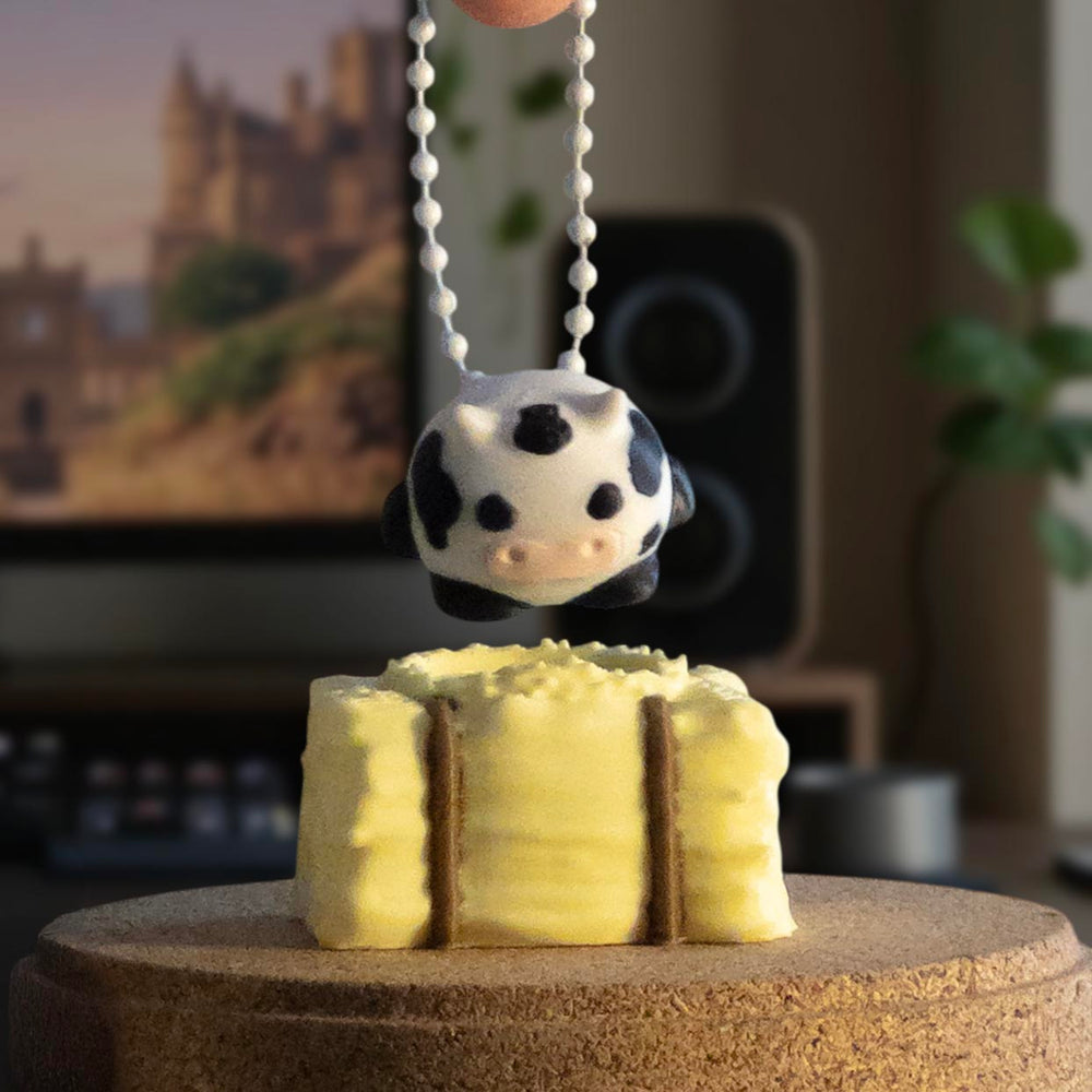 A black and white spotted cow charm being held by a keychain above a stack of yellow hay bales on a tabletop.
