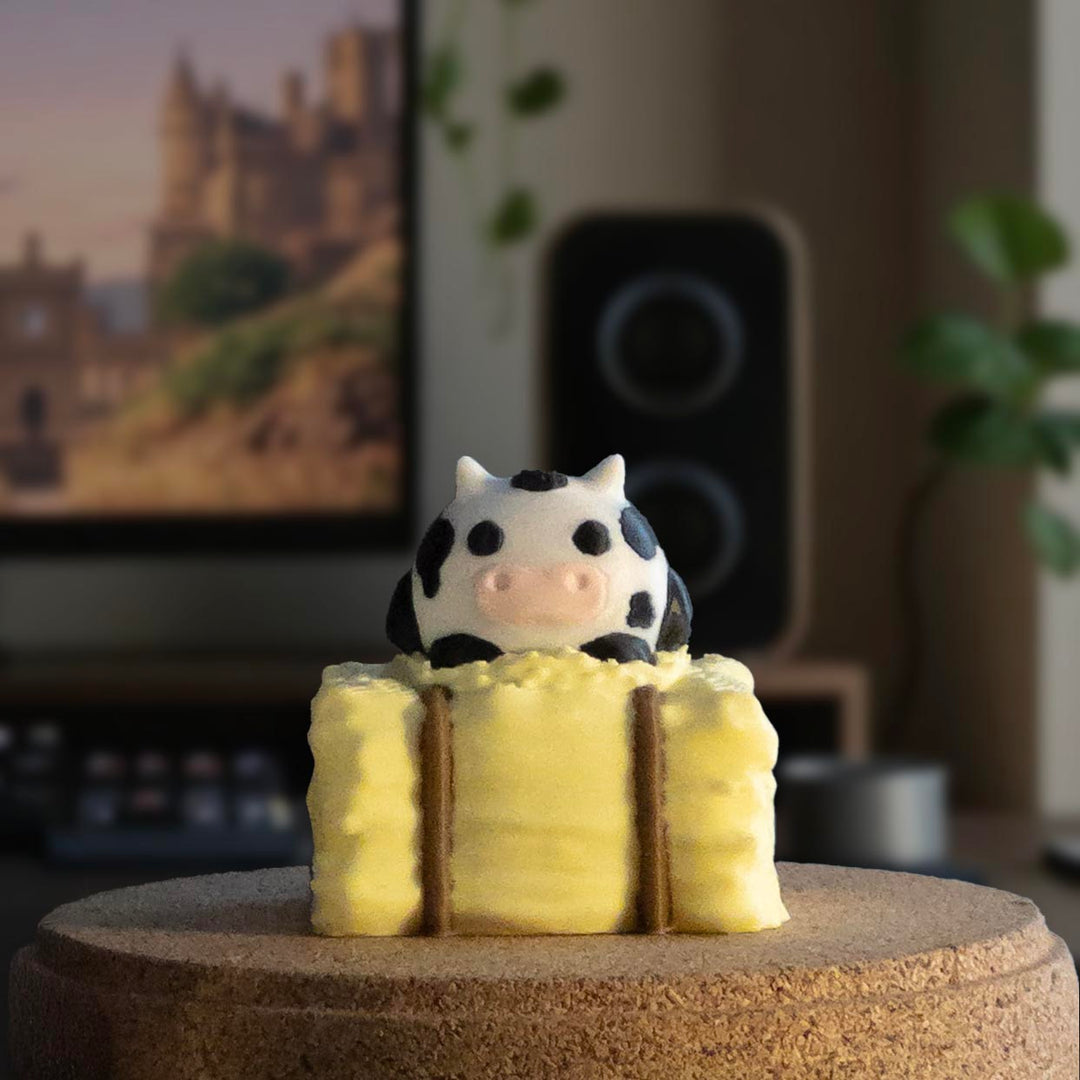 A cute black and white spotted cow charm sitting on top of a stack of yellow hay bales, displayed on a tabletop.