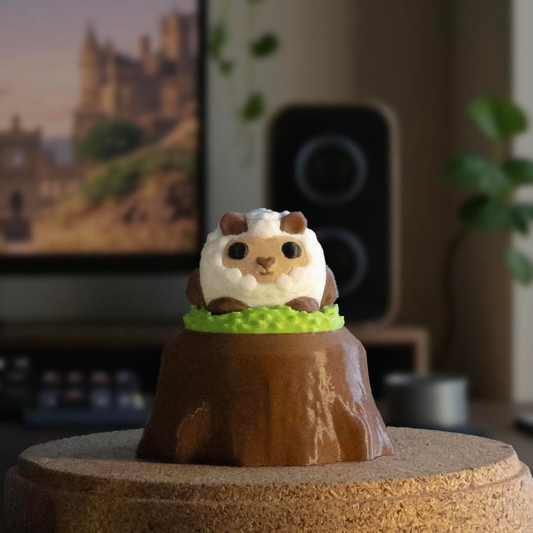 A fluffy sheep charm with brown ears and legs sitting on a grassy patch atop a tree stump display, placed on a tabletop.